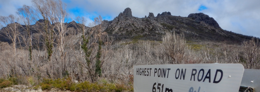 The sign which marks the start of the walk. It reads "highest point on road. 651m annual rain fall. 190cm" The rocky spires that make up the needles are foreboding in the background, cast in shadow.